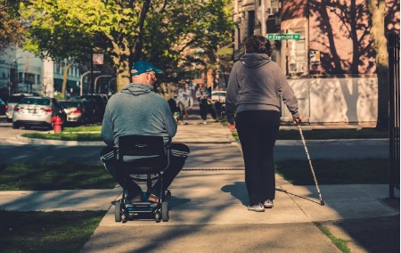 A person riding a scooter next to a person with a cane.